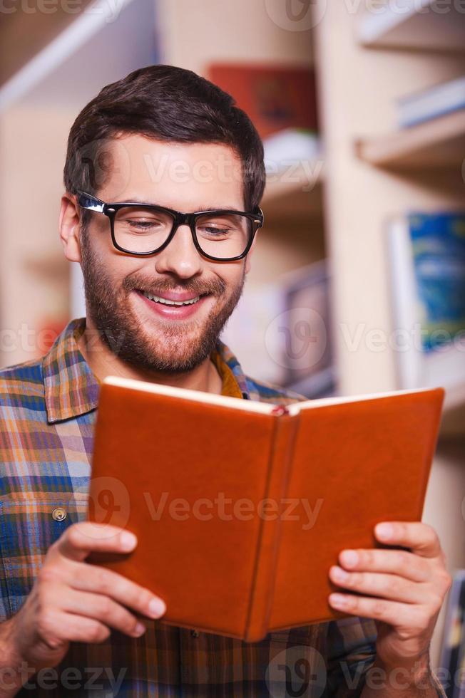 Reading his favorite book. Handsome young man reading book and smiling while standing in front of the bookshelf photo