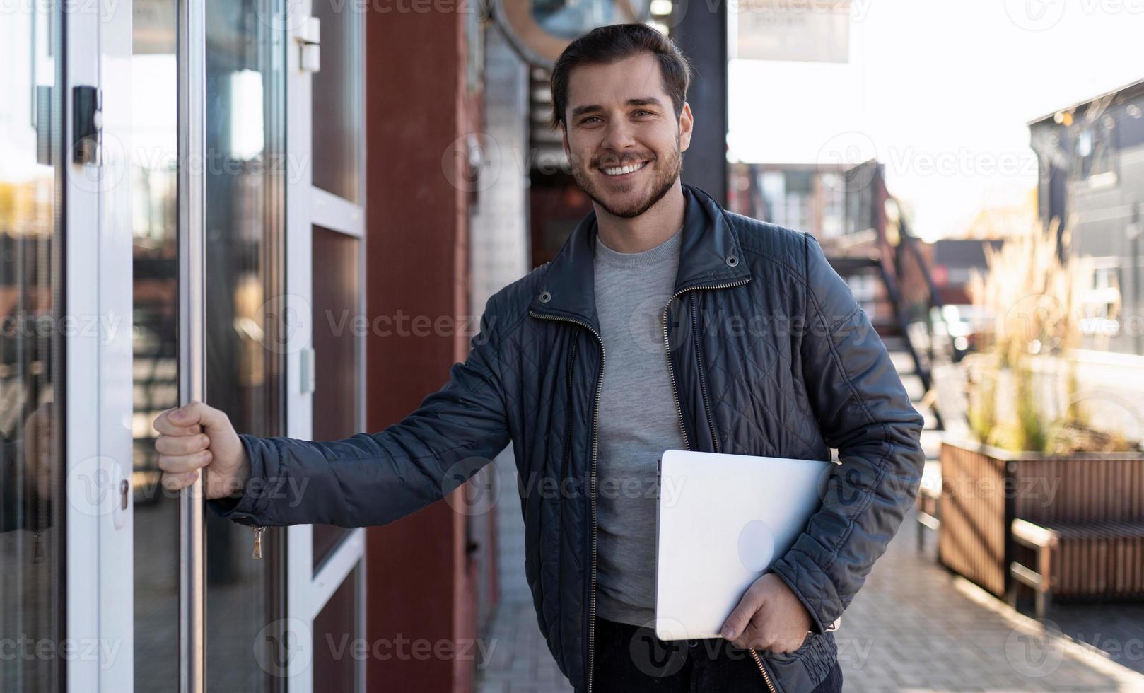 a male IT specialist with a laptop in his hands enters the office building with a wide smile on his face photo