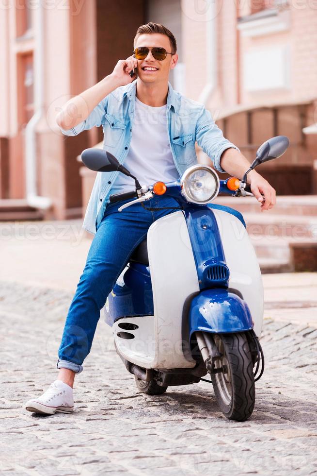 Inviting friends for a ride. Cheerful young man sitting on scooter and talking on the mobile phone photo