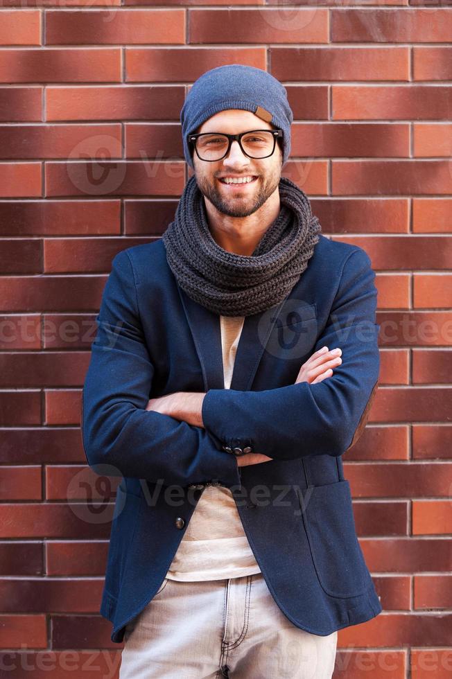 Confident in his perfect style. Handsome young man in smart casual wear keeping arms crossed and smiling while leaning at the brick wall photo