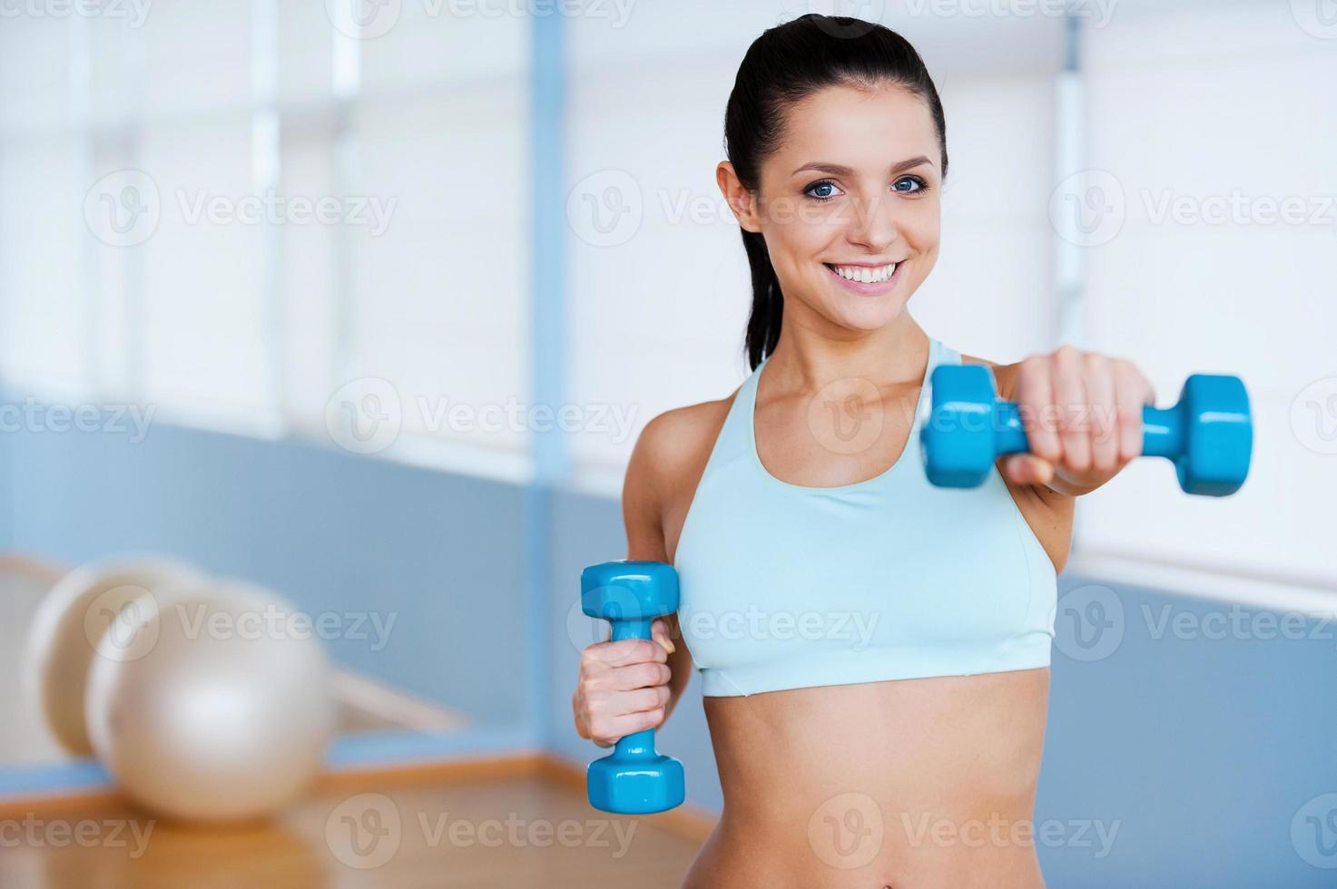 Exercising with dumbbells. Beautiful young woman in sports clothing exercising with dumbbells and smiling while standing in health club photo