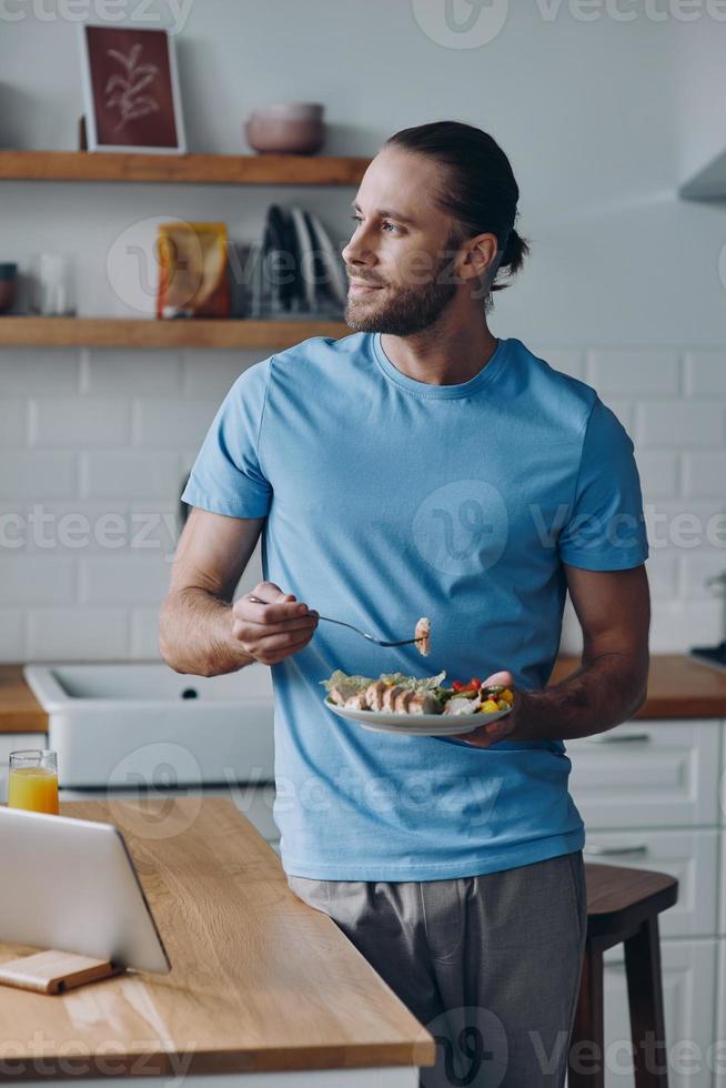Dreamful young man enjoying lunch while leaning at the kitchen counter at home photo