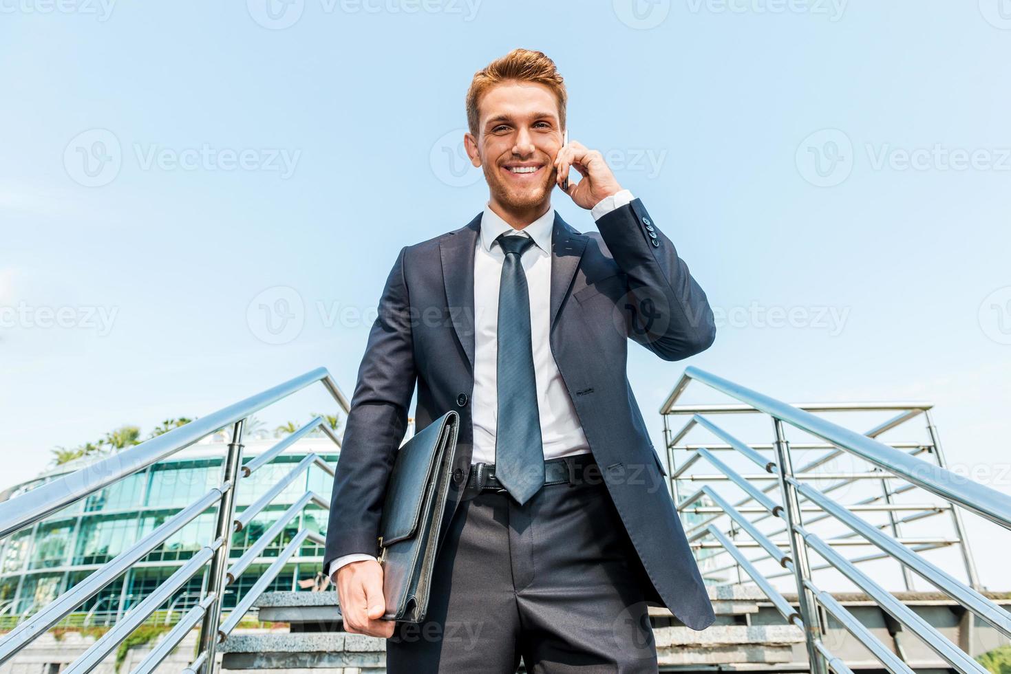 Businessman on the phone. Low angle view of confident young man in formalwear talking on the mobile phone and smiling while moving down by staircase photo