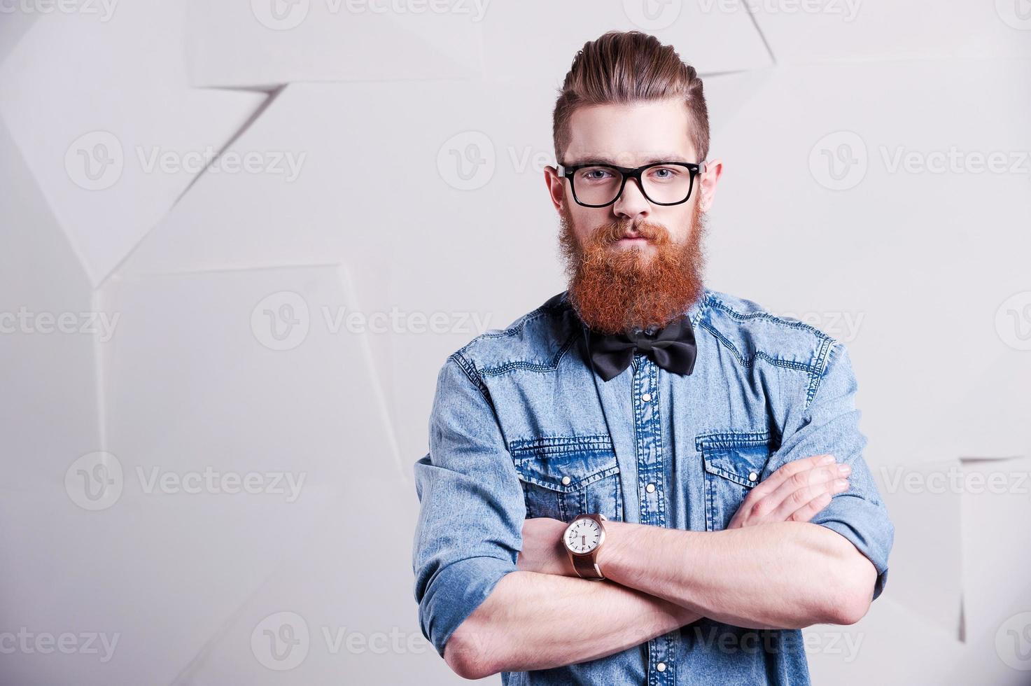 Looking trendy. Handsome young bearded man in glasses keeping arms crossed and looking at camera photo