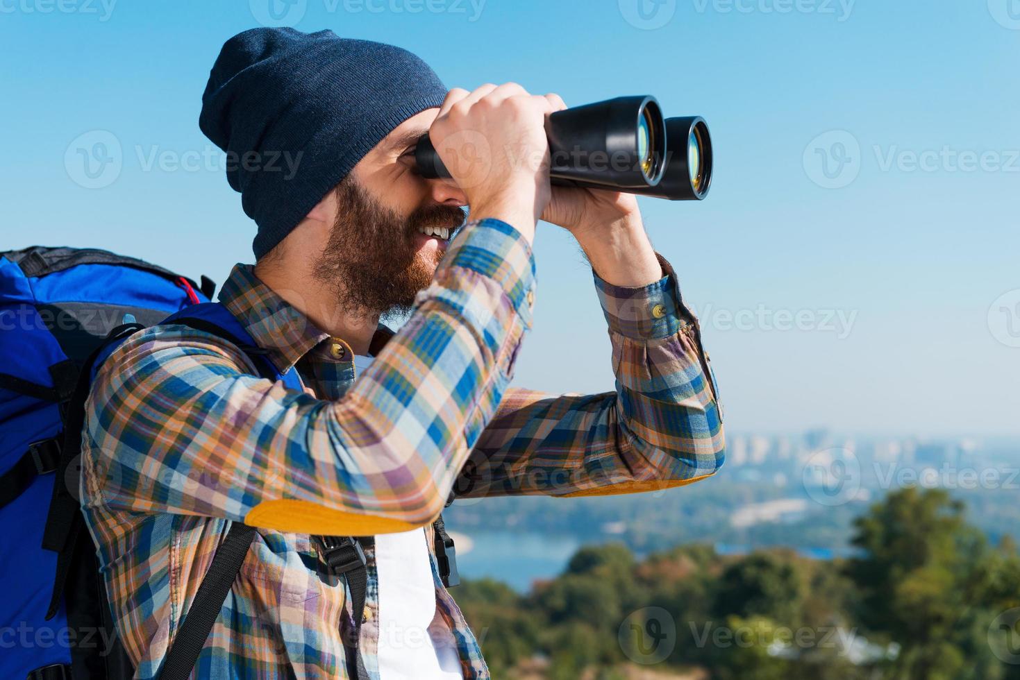 feliz de ser un explorador. apuesto joven barbudo llevando mochila y mirando a través de binoculares con una sonrisa foto