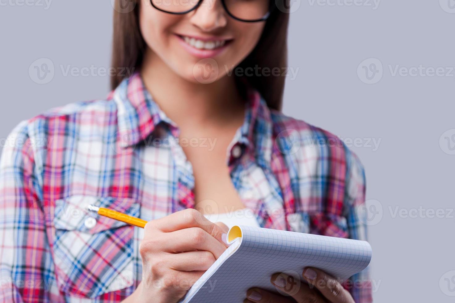 Making notes. Close-up of young women writing in notebook by a pencil while standing against grey background photo