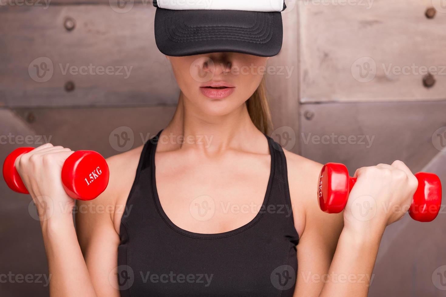 Strong and charming. Muscular woman holding dumbbells and wearing cap  while standing against metal background photo