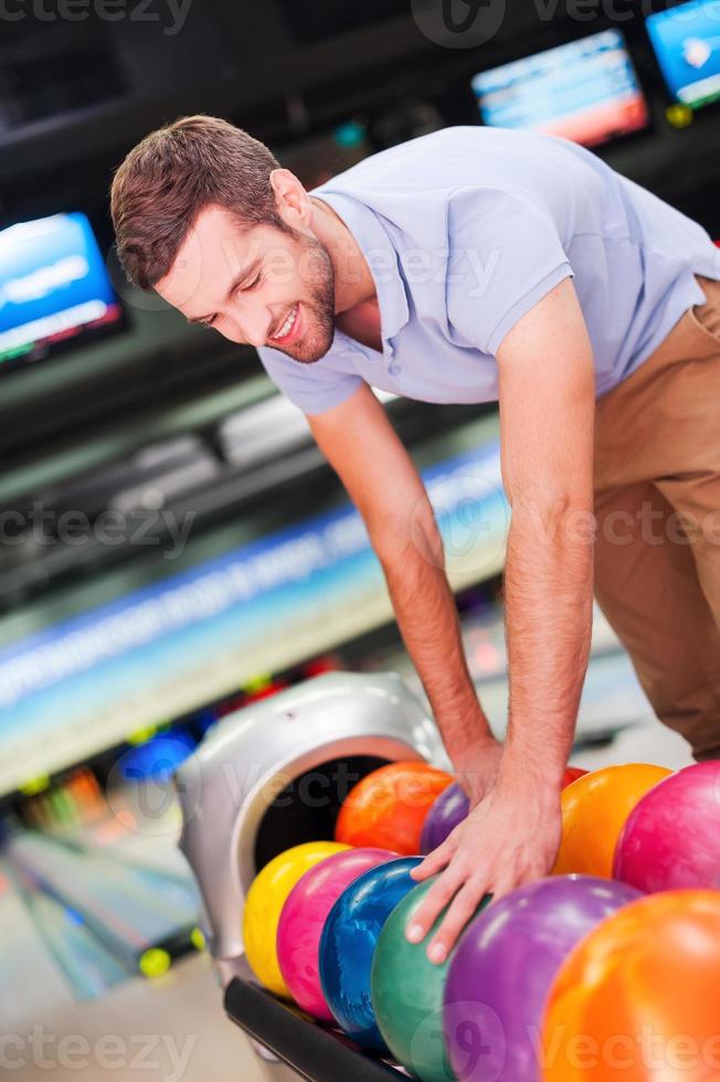 eligiendo la bola de la suerte. un joven alegre eligiendo una bola de bolos y sonriendo mientras se enfrenta a las boleras foto