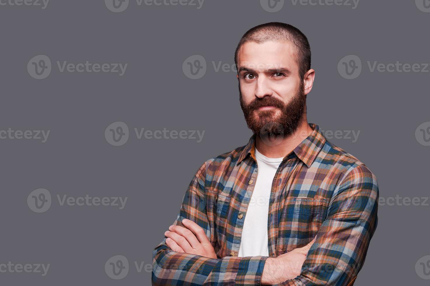 In friends with irony. Handsome young bearded man keeping arms crossed and expressing ironic smile while standing against grey background photo