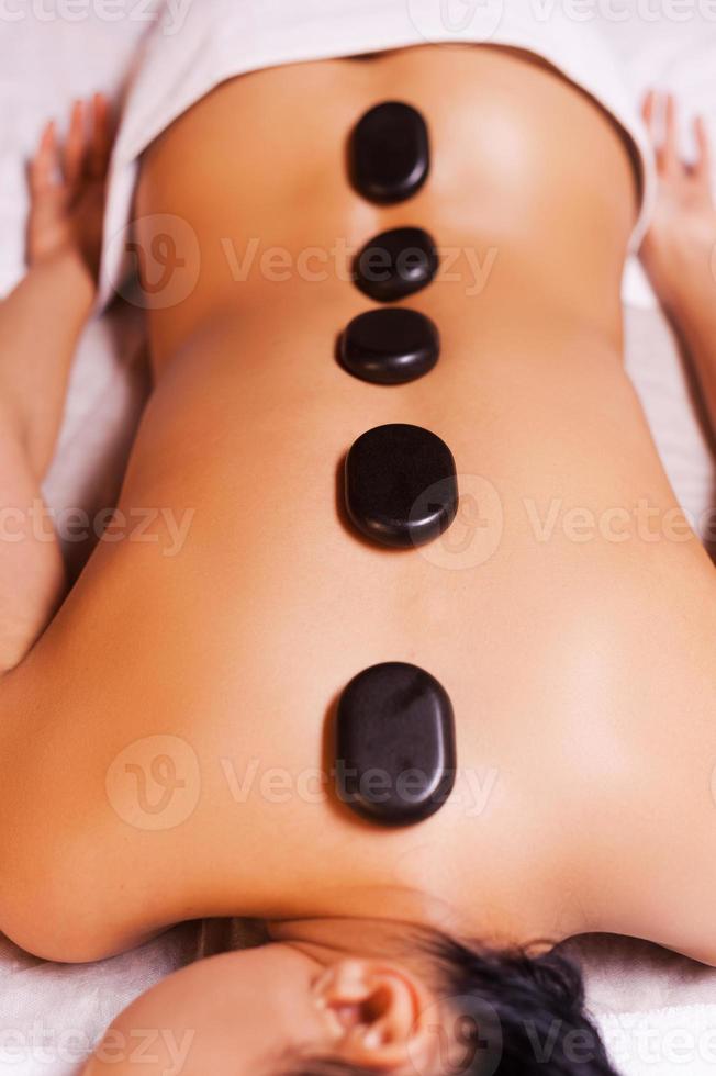 Hot stone therapy. Top view of young woman with shiny skin lying on front with spa stones on her back photo