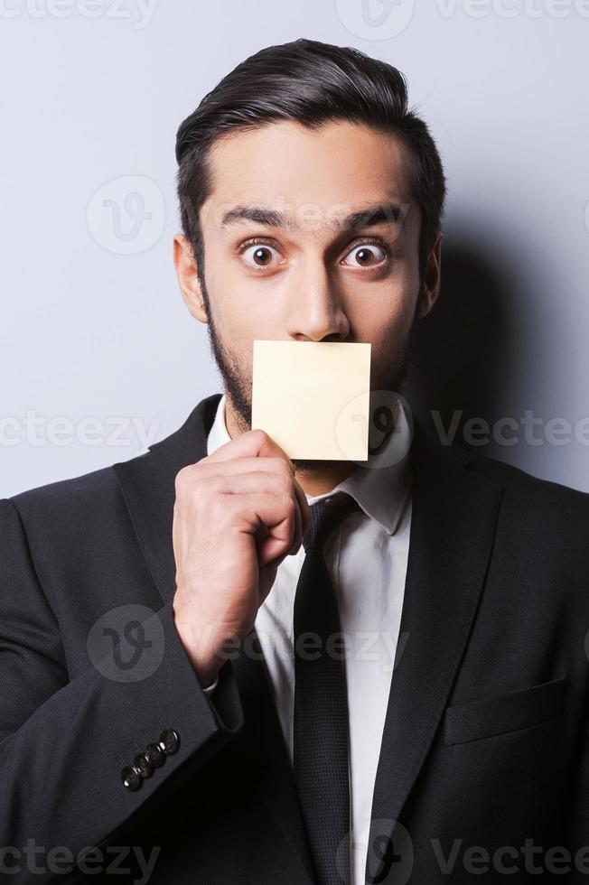 With humor to the deals. Portrait of surprised young man in formalwear covering his mouth with the sticky note while standing against grey background photo