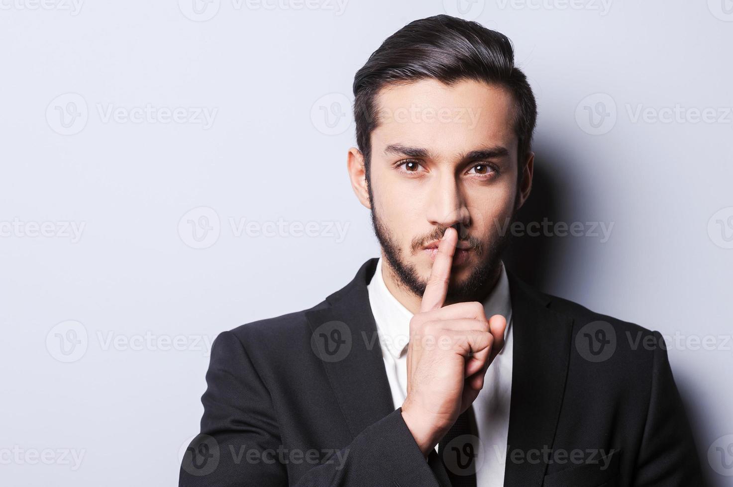 Keep my secret  Serious young man in formalwear holding finger on lips and looking at camera while standing against grey background photo