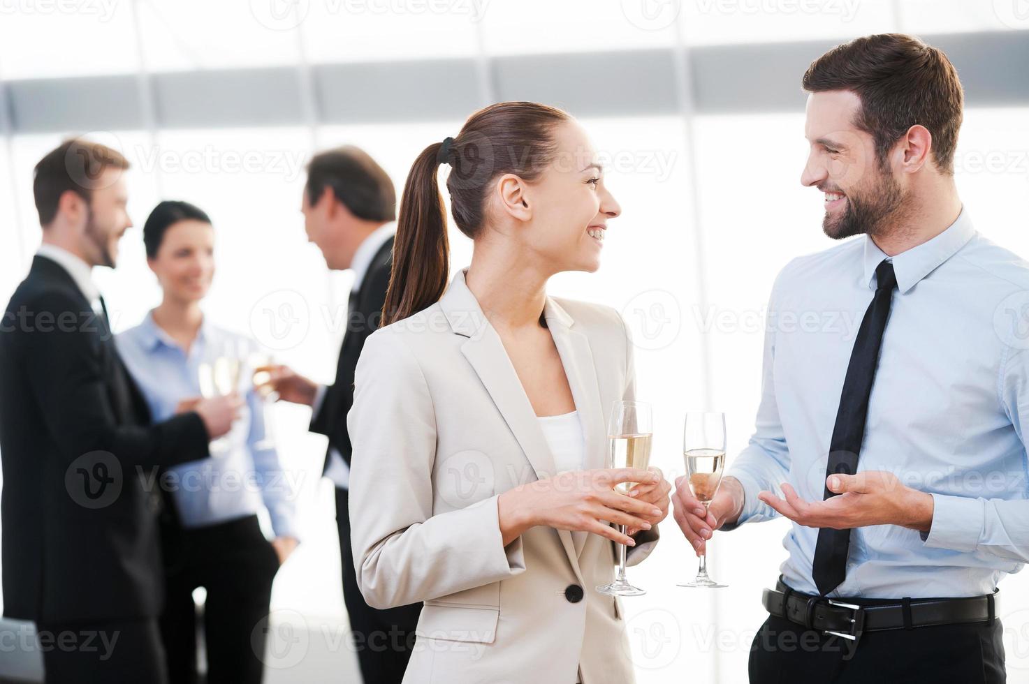 Celebrating their common success. Two cheerful business people drinking champagne and talking while other people communicating in the background photo