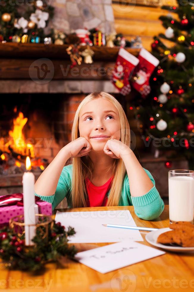 What do I wish Cute little girl day dreaming while sitting at home with Christmas tree and fireplace in the background photo