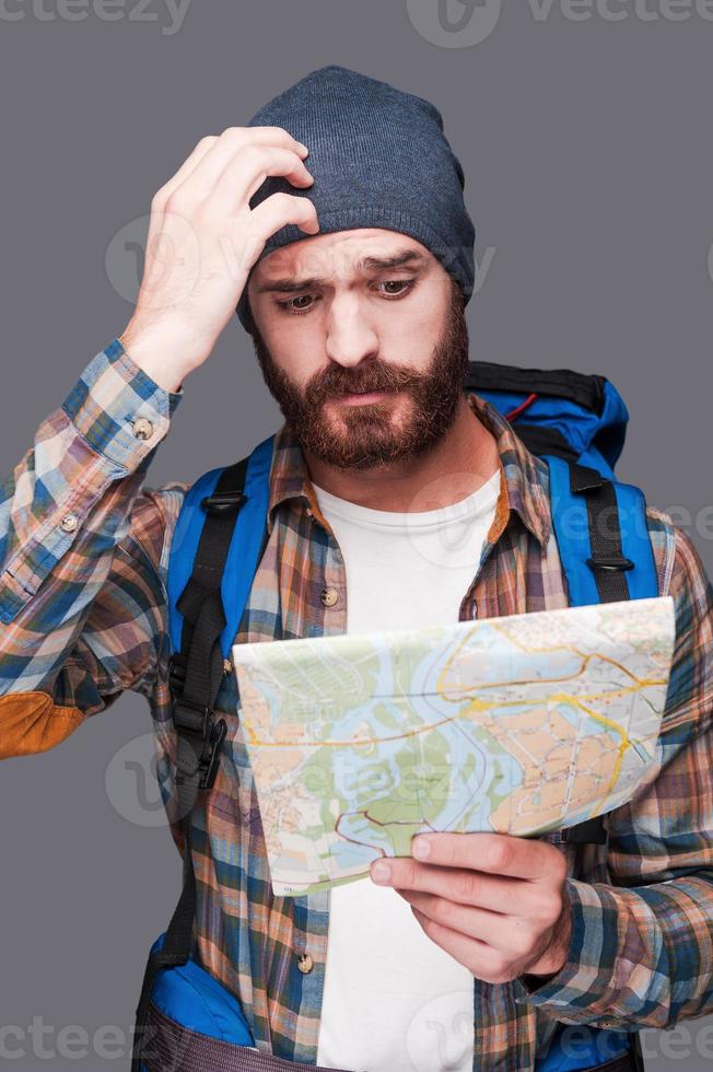 Lost in somewhere. Frustrated young bearded man carrying backpack and examining map while scratching head and standing against grey background photo