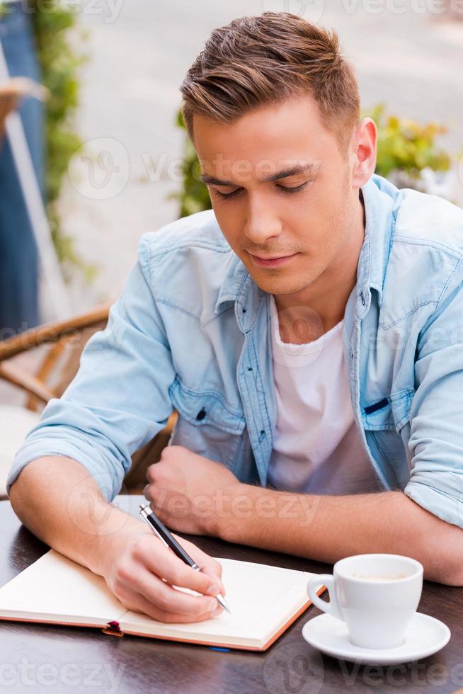 Just inspired. Confident young man writing something in note pad while sitting in sidewalk cafe photo