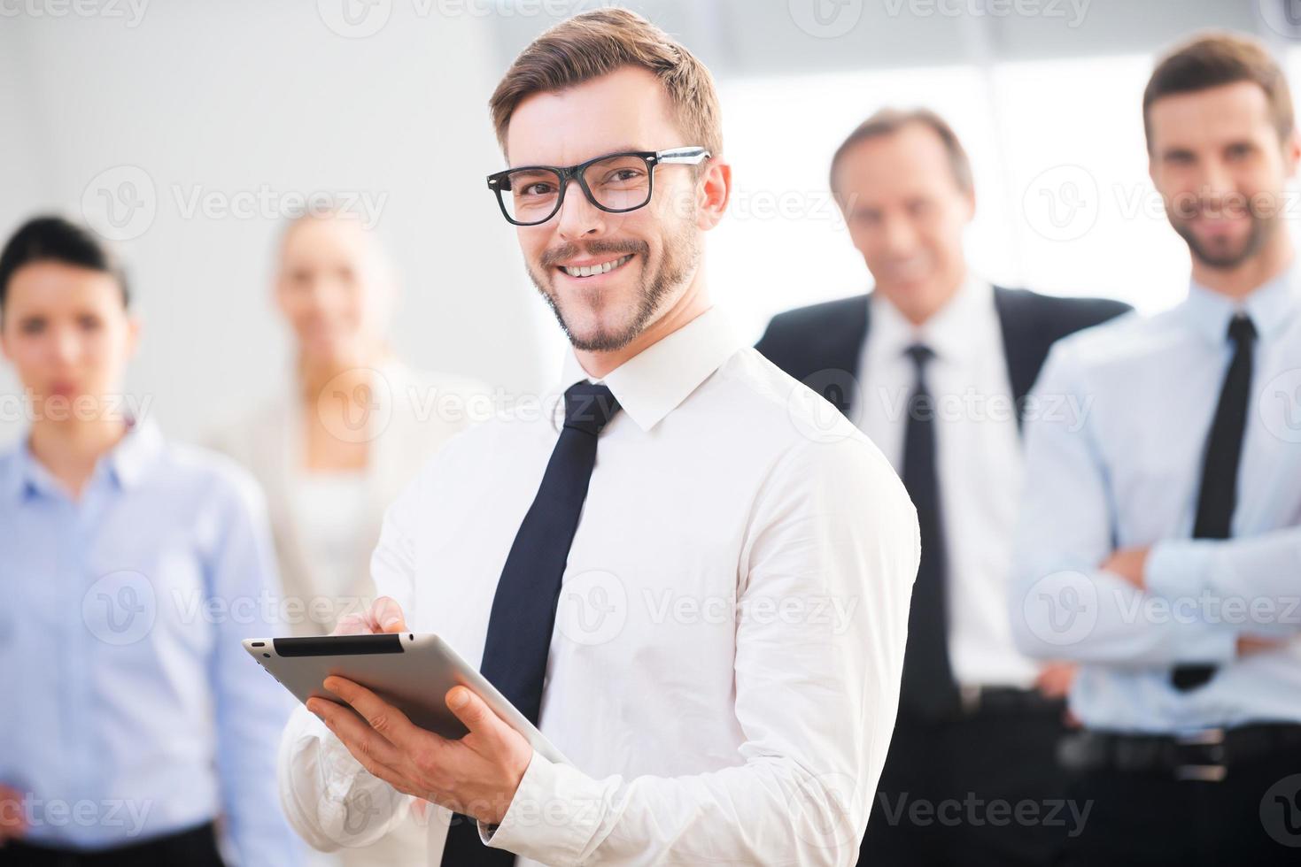 experto en negocios confiado. un joven hombre de negocios confiado sosteniendo una tableta digital y sonriendo mientras sus colegas estaban parados en el fondo foto