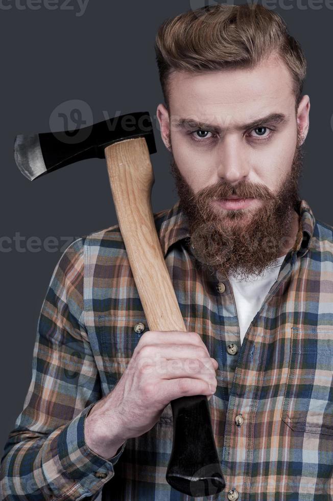 Just me and my axe. Confident young bearded man carrying a big axe on shoulder and looking at camera while standing against grey background photo
