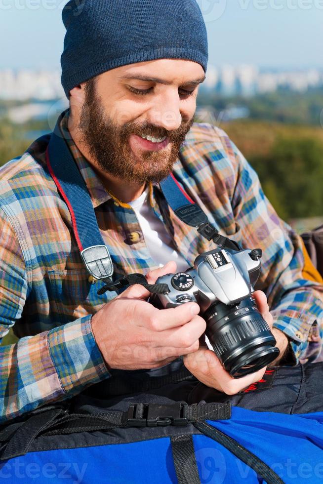 recordando todo el viaje. apuesto joven sentado cerca de la mochila y sosteniendo la cámara foto