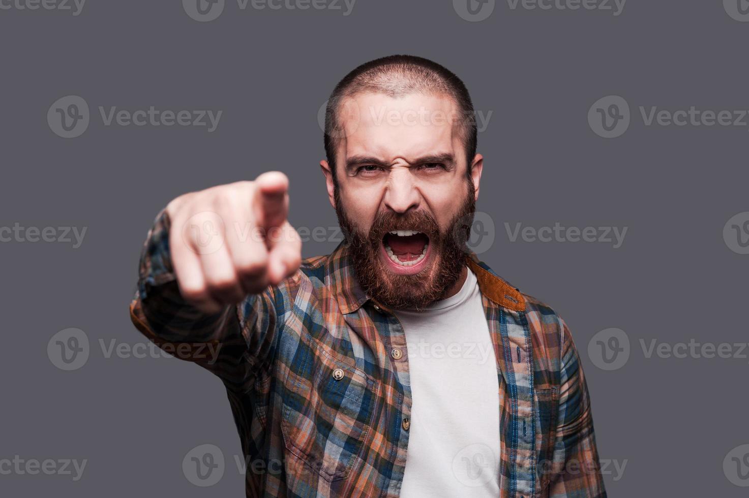 Hey you Furious young bearded man pointing you and shouting while standing against grey background photo