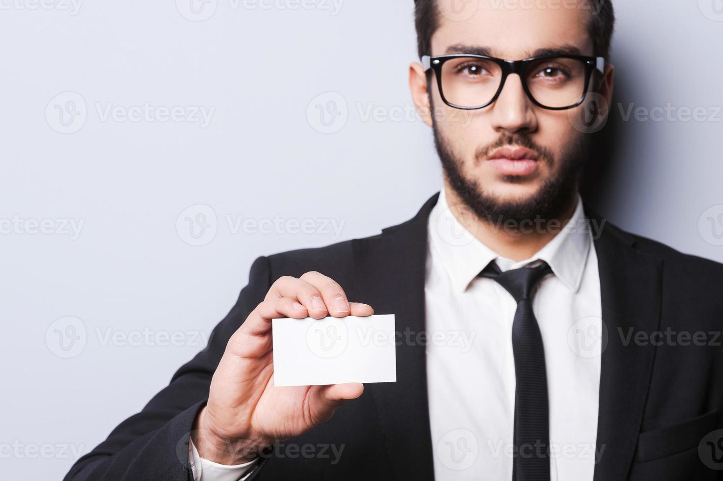 Llame a este número apuesto joven en ropa formal estirando una tarjeta de visita mientras está de pie contra un fondo gris foto