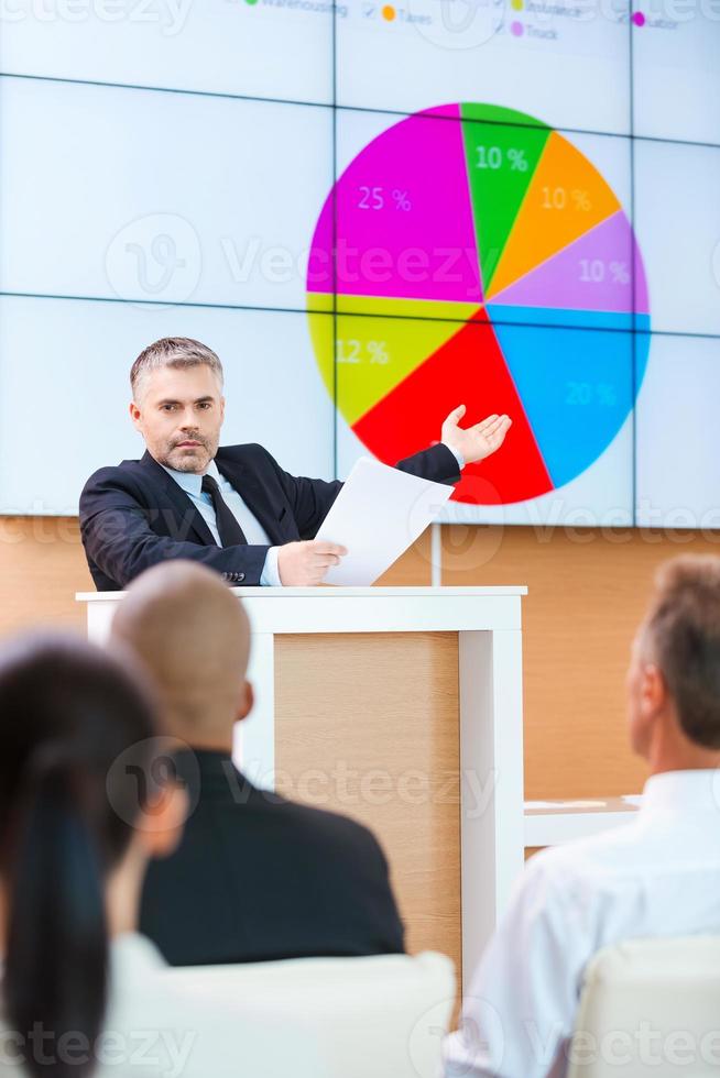 Public speaker. Confident grey hair man in formalwear pointing projection screen with graph on it while making presentation in conference hall with people on the foreground photo