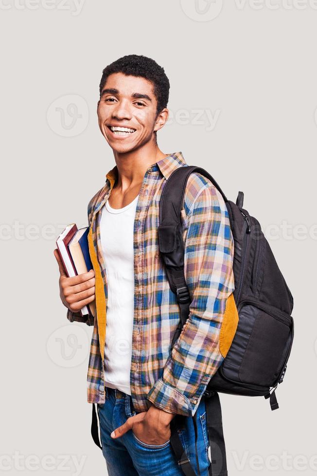 Me encanta estudiar. apuesto joven estudiante afroamericano sosteniendo libros y sonriendo mientras está de pie contra un fondo gris foto