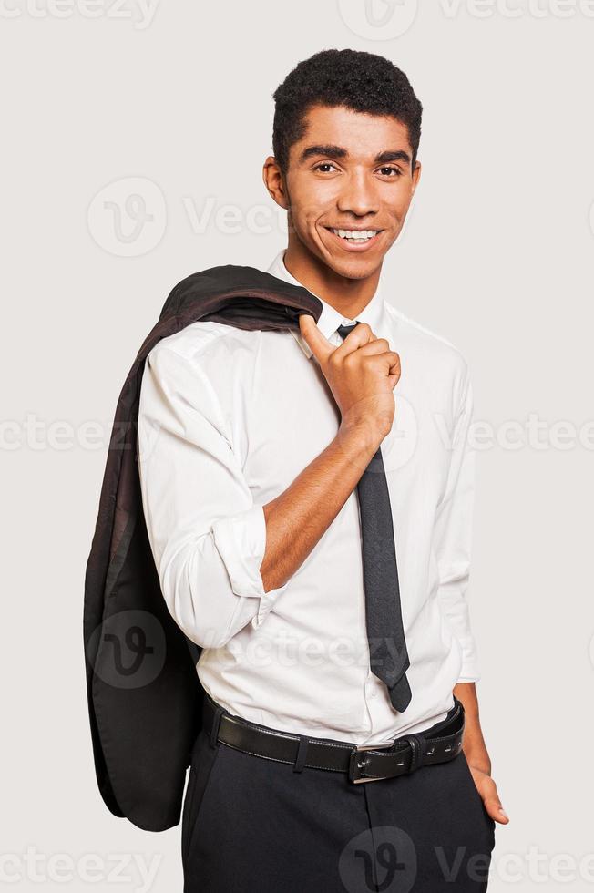 un joven afroamericano confiado vestido con ropa formal que lleva su chaqueta sobre los hombros y mira la cámara mientras se enfrenta a un fondo gris foto