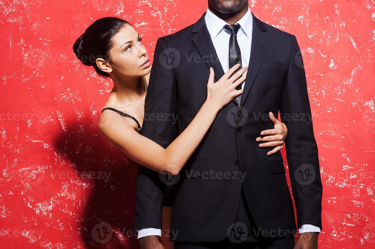 Passion and desire. Cropped image of man in formalwear standing against red background while woman embracing him from behind photo