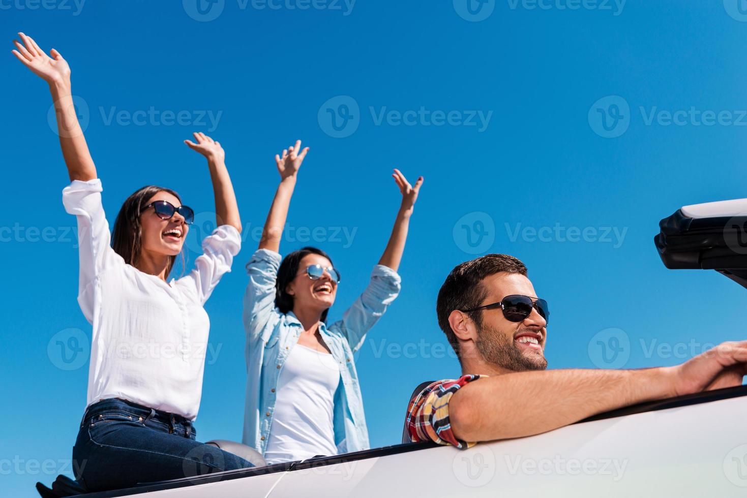 Enjoying road trip. Three young happy people enjoying road trip in their white convertible while two beautiful woman raising arms and smiling photo