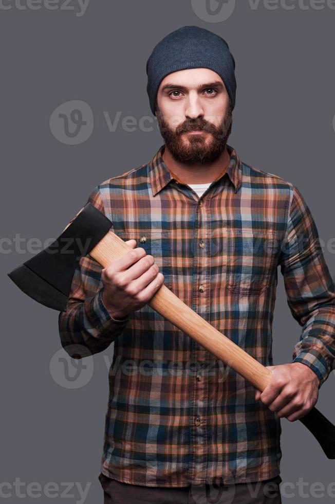 Confident lumberjack. Confident young bearded man holding a big axe and looking at camera while standing against grey background photo