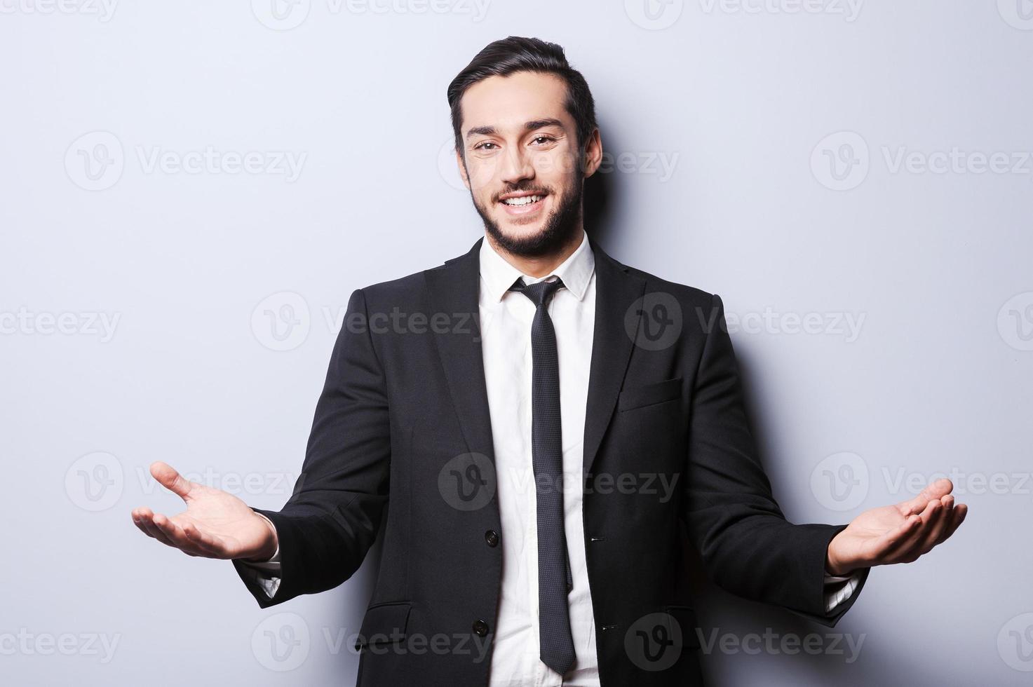 You are welcome Cheerful young man in formalwear looking at camera and gesturing while standing against grey background photo