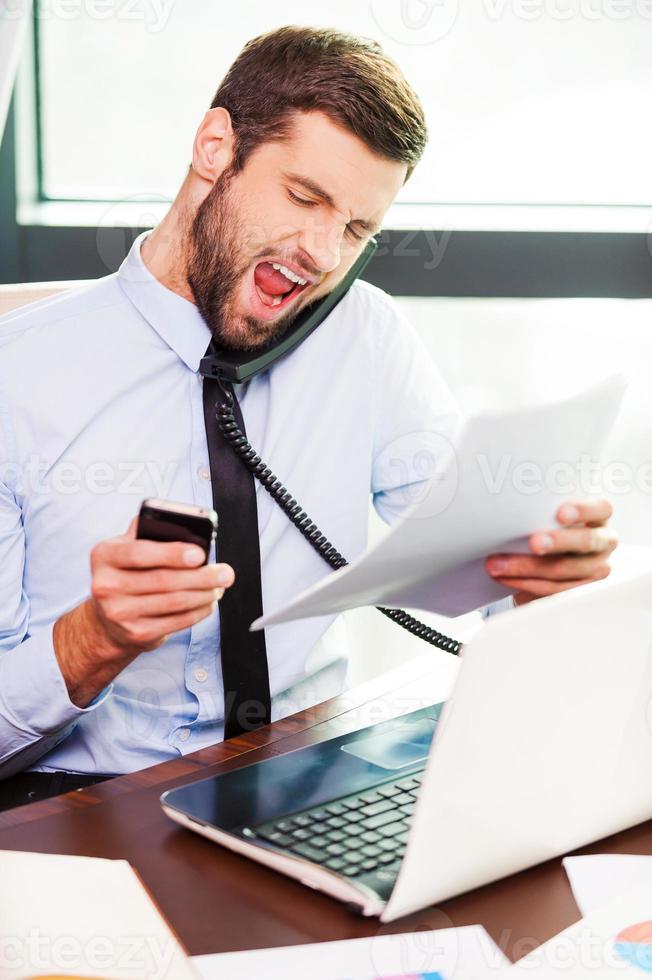 Working under pressure. Furious young man in shirt and tie talking on the phone and shouting while holding documents and mobile phone in his hands photo