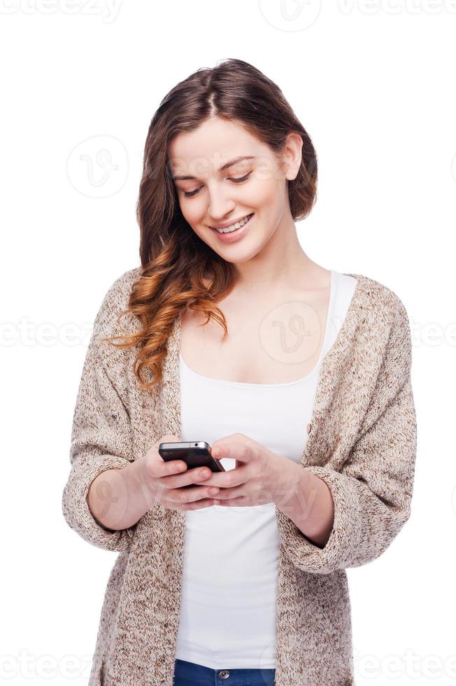 Typing message to friend. Cheerful young woman holding mobile phone and smiling while standing isolated on white photo