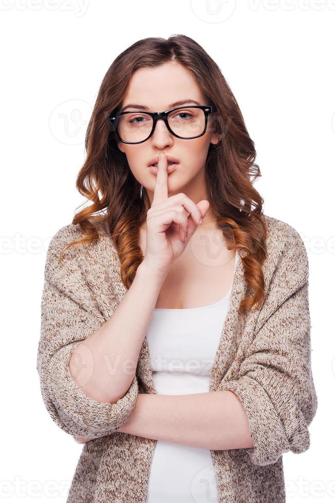 mantenga el silencio mujer joven atractiva en anteojos mirando a la cámara y sosteniendo el dedo en los labios mientras está de pie aislado en blanco foto