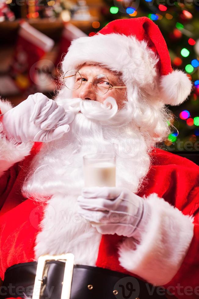 me encanta la leche alegre santa claus sosteniendo un vaso con leche y ajustando su bigote con el árbol de navidad en el fondo foto