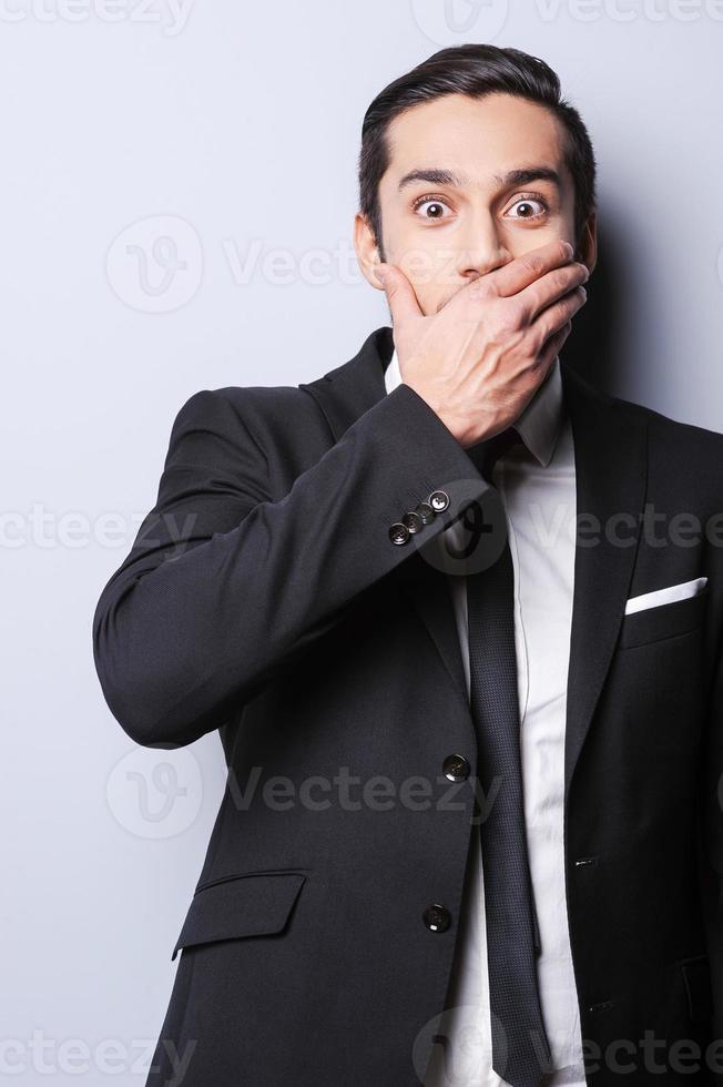 Hope, I do not say too much. Excited young man in formalwear covering mouth with hand and looking at camera while standing against grey background photo