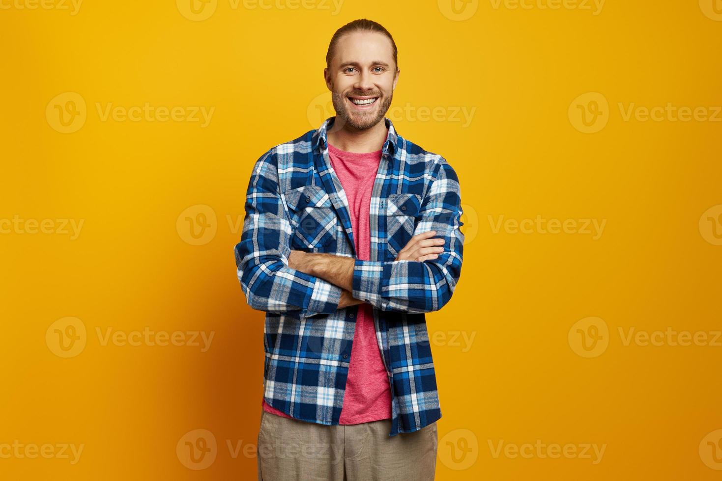 Cheerful young man keeping arms crossed while standing against yellow background photo