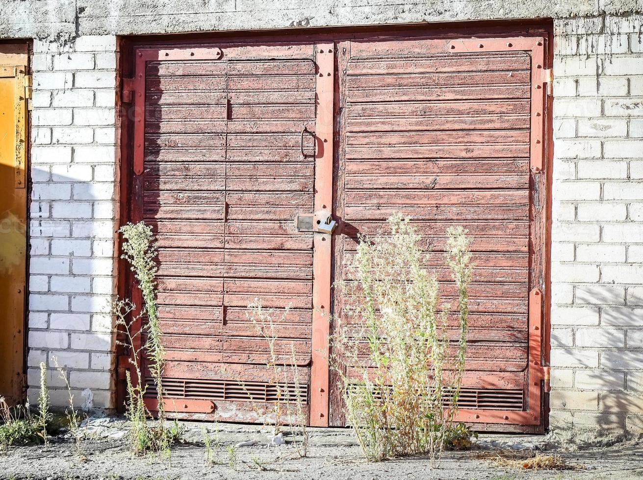 Wide brown wooden door locked with a padlock with grass growing in front of the entrance with midday shadows photo