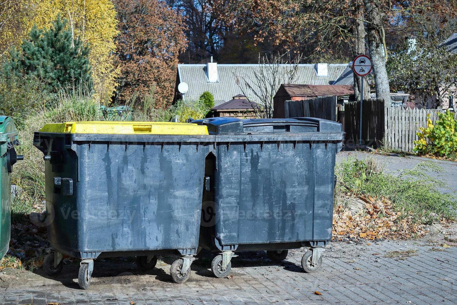 Two black garbage containers on wheels standing on pathway in autumn partk with visible house photo