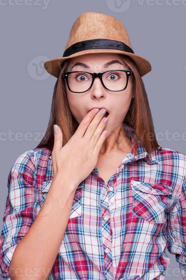 It can not be truth Surprised young woman in glasses and funky hat covering mouth with hand and looking at camera while standing against grey background photo