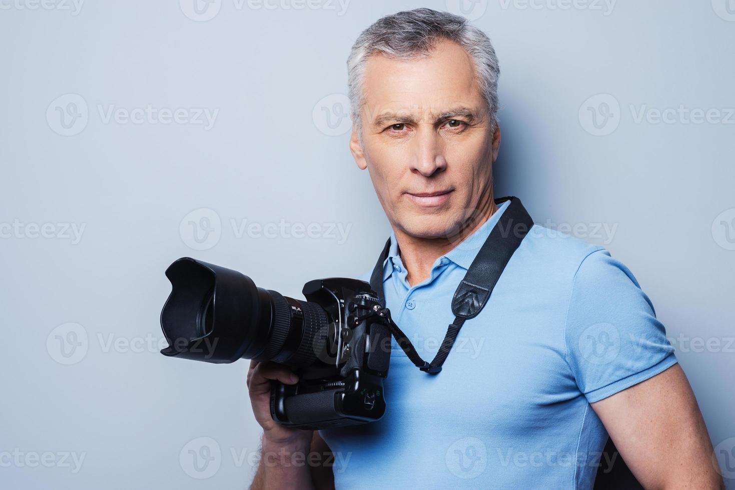 fotógrafo profesional. retrato de un hombre maduro confiado en una camiseta sosteniendo una cámara mientras se enfrenta a un fondo gris foto