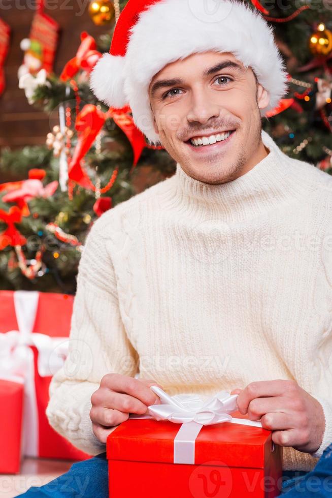 My Christmas present. Handsome young man in Santa hat opening gift box and smiling with Christmas Tree in the background photo