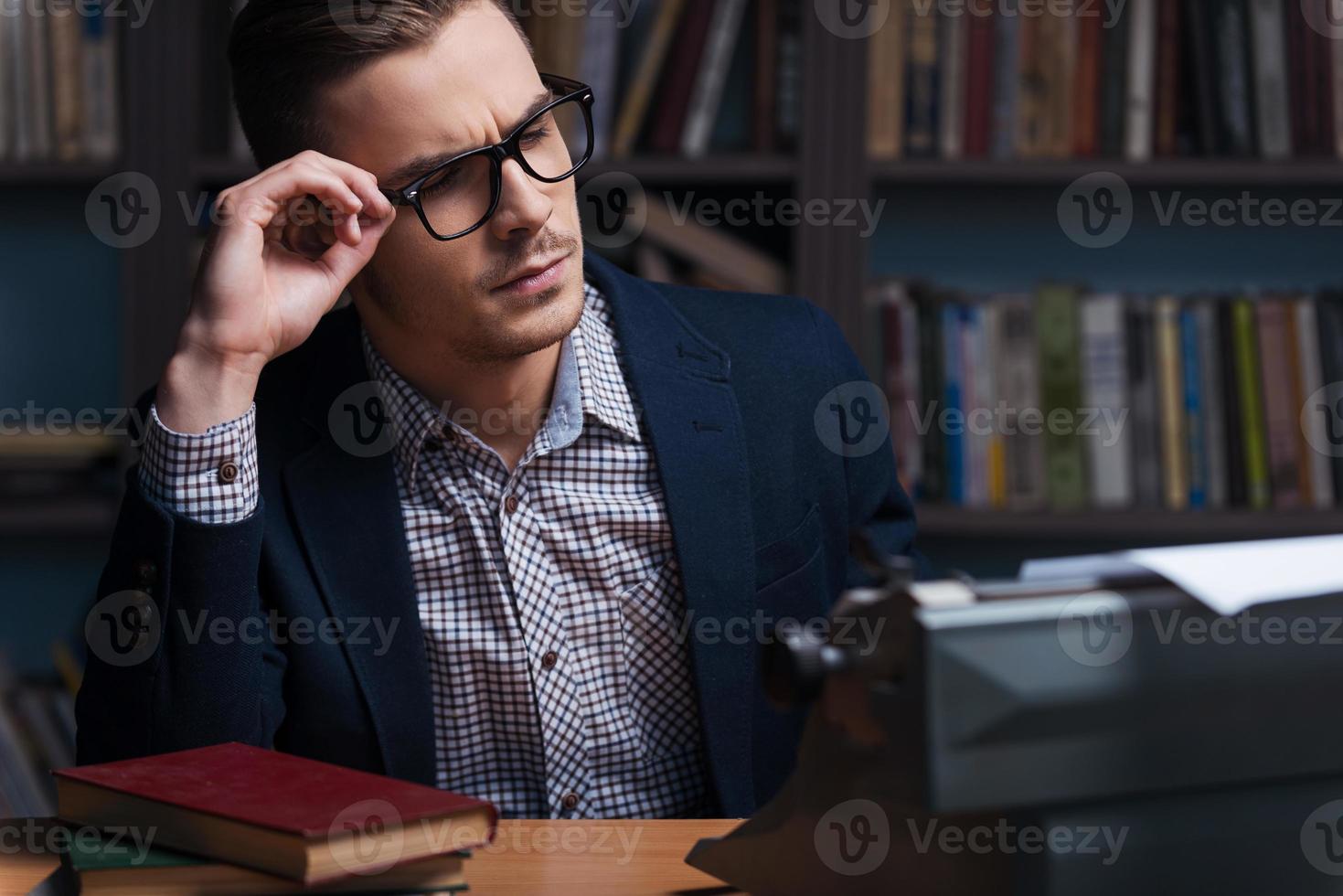 Thoughtful author. Thoughtful young author working at the typewriter and adjusting his eyeglasses while sitting at his working place with bookshelf in the background photo