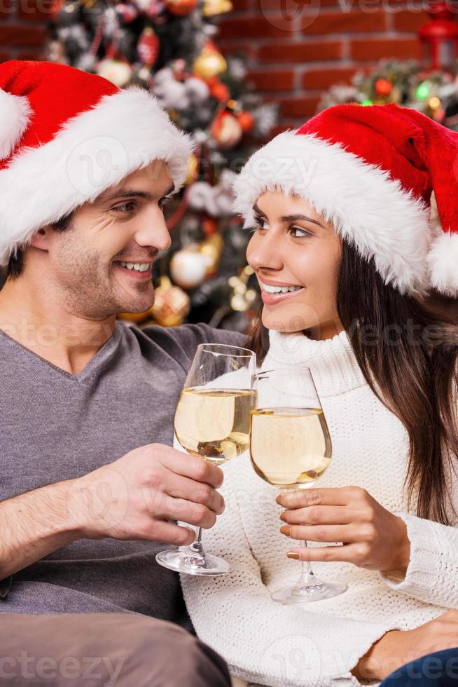 feliz año nuevo feliz joven pareja amorosa con sombreros de santa uniéndose entre sí y sosteniendo copas de vino con un árbol de navidad en el fondo foto