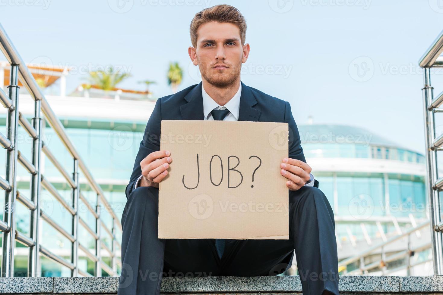 Looking for a job. Handsome young man in formalwear holding poster with job text message while sitting outdoors and with building structure in the background photo