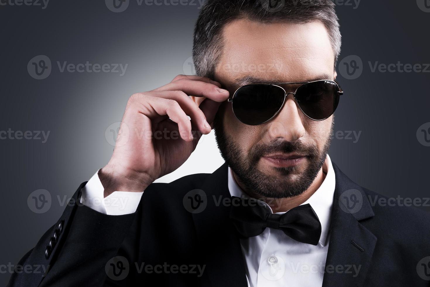 Confident heartbreaker. Portrait of handsome mature man in formalwear adjusting his sunglasses while standing against grey background photo