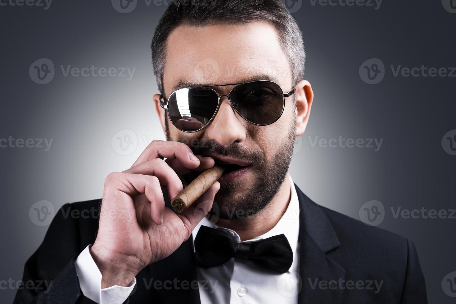 Confident and bossy. Portrait of handsome mature man in formalwear and sunglasses smoking cigar while standing against grey background photo