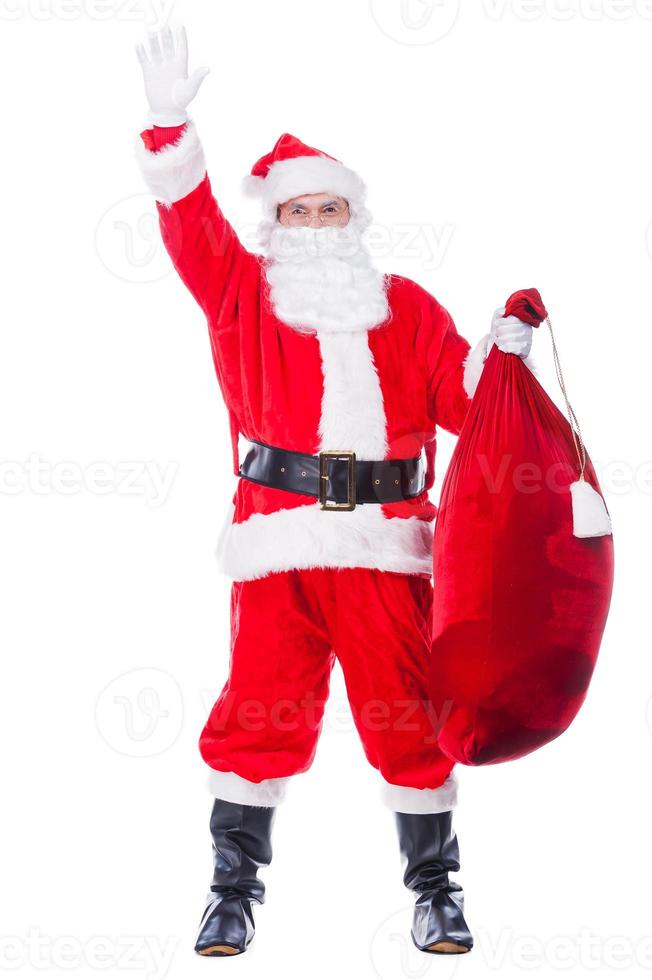 Merry Christmas and Happy New Year Full length of Traditional Santa Claus carrying sack with presents and waving to you while standing against white background photo