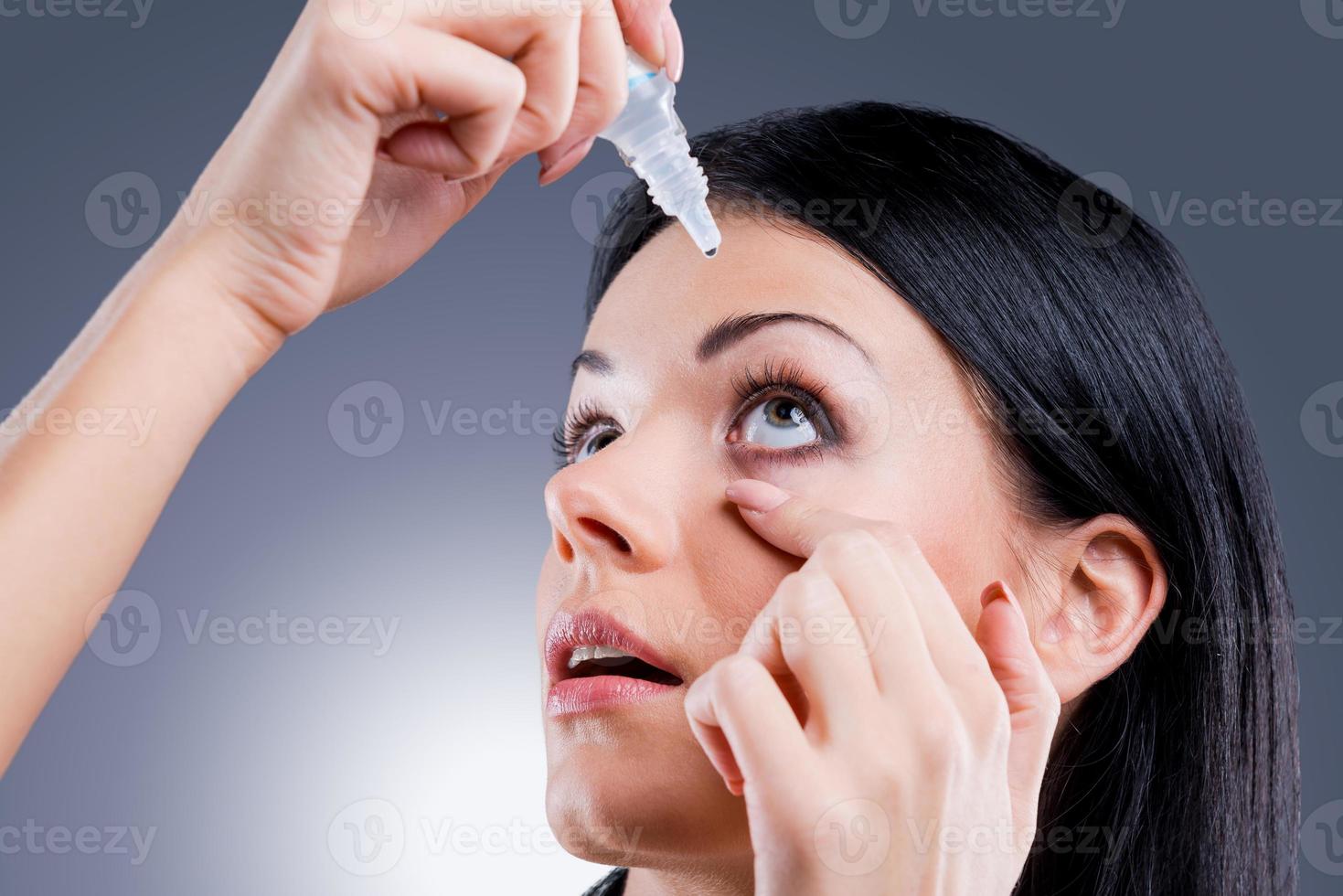 Taking care of her vision. Side view of young women applying eye drops while standing against grey background photo